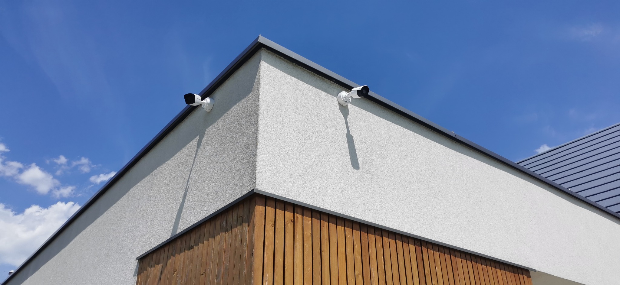 Dwie białe kamery monitoringu zamontowane na rogu budynku z jasną elewacją i drewnianą okładziną, widok z dołu na tle błękitnego nieba.