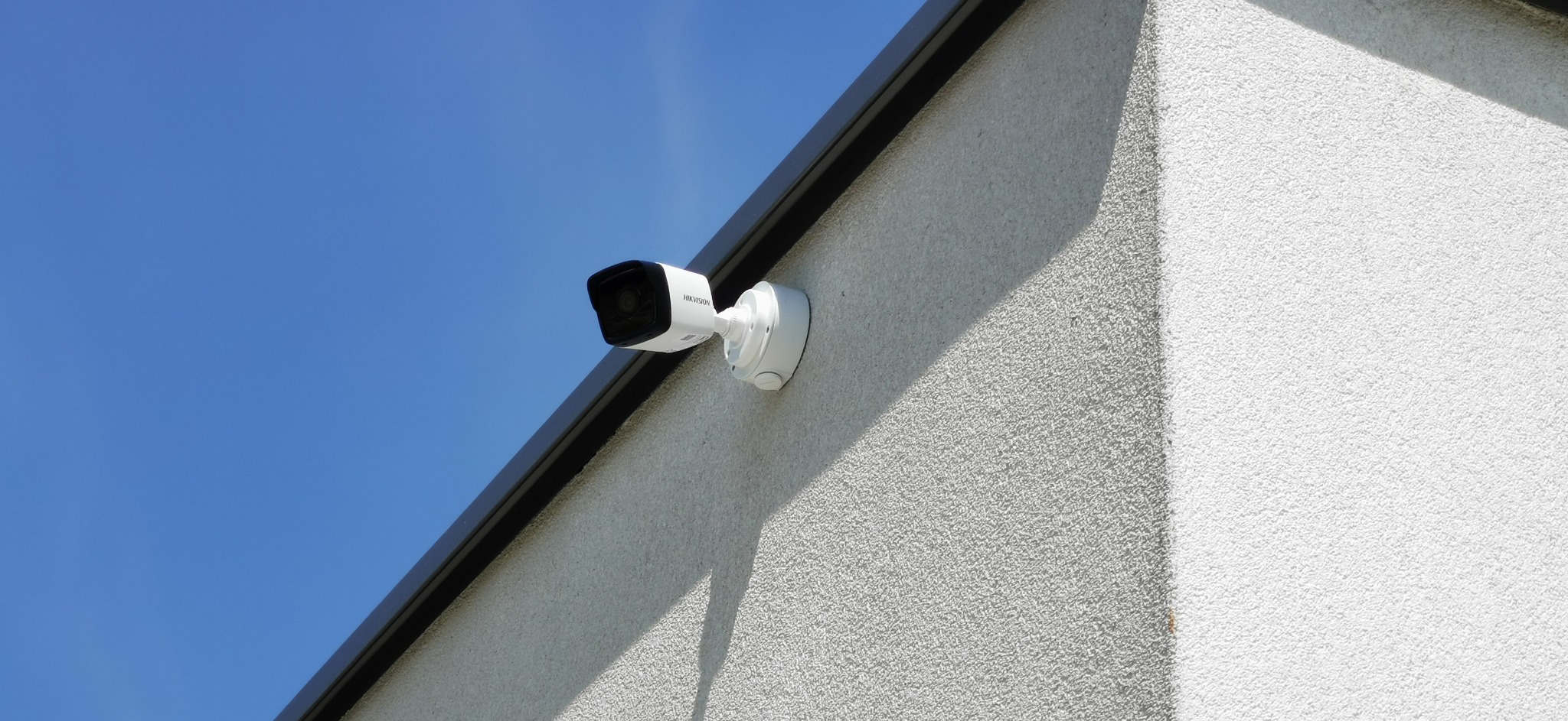 Biała kamera monitoringu Hikvision zamontowana na teksturowanej, jasnoszarej ścianie budynku pod błękitnym niebem. Widoczny fragment czarnej rynny dachowej.