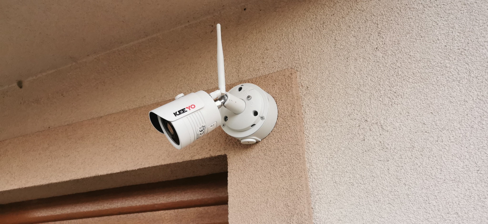 Zewnętrzna kamera monitoringu marki KEEYO z anteną, zamontowana na chropowatej ścianie budynku o dwóch odcieniach beżu, widoczna z boku.