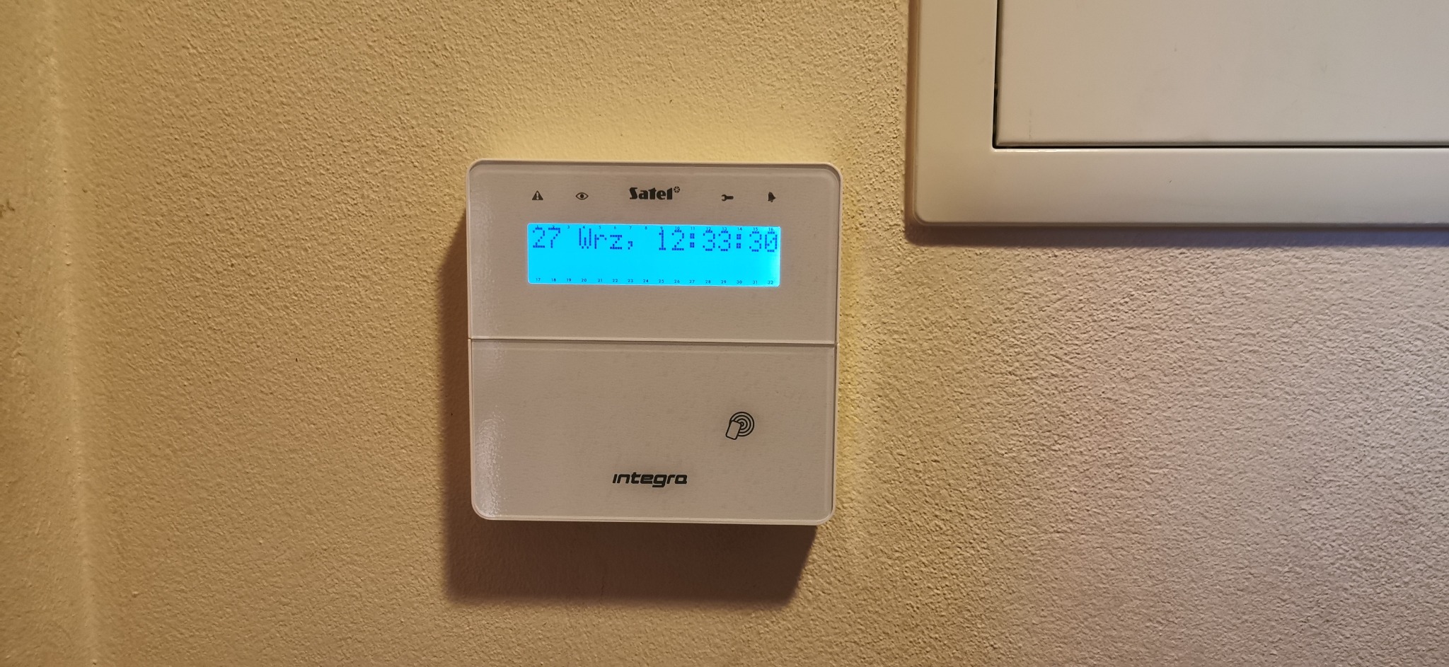 Biały panel sterowania alarmem Satel Integra zamontowany na beżowej ścianie z widocznym wyświetlaczem LCD pokazującym datę i godzinę.