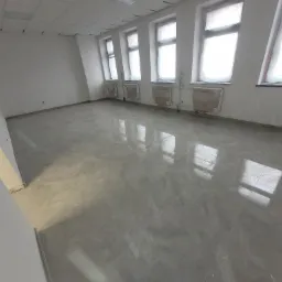 Wykończone wnętrze z szarymi płytkami podłogowymi, okna zabezpieczone folią malarską, grzejniki zakryte, sufit podwieszany.