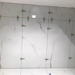 Ściana wyłożona białymi płytkami imitującymi marmur, z widocznymi zielonymi krzyżykami dystansowymi i czarnymi liniami fug, w trakcie prac wykończeniowych.