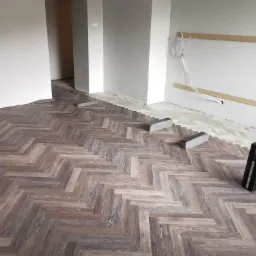 Fragment podłogi w jodełkę, ułożonej z paneli laminowanych w odcieniach brązu i szarości, z widocznymi akcesoriami montażowymi i paczką paneli PROJECT FLOORING w tle, w trakcie prac wykończeniowych.