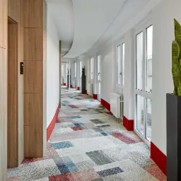 Długi korytarz w budynku biurowym z wykładziną dywanową w geometryczne wzory, białymi ścianami, grzejnikami pod oknami i rośliną w czarnej donicy.