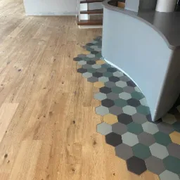 Jasny parkiet drewniany przechodzi w mozaikę heksagonalnych płytek w odcieniach szarości, zieleni i żółci, otaczających szarą zabudowę meblową; w tle widoczne schody.