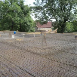 Siatka zbrojeniowa na przygotowanej powierzchni pod wylewkę, widoczne wystające pręty, w tle zieleń drzew i fragment budynku.