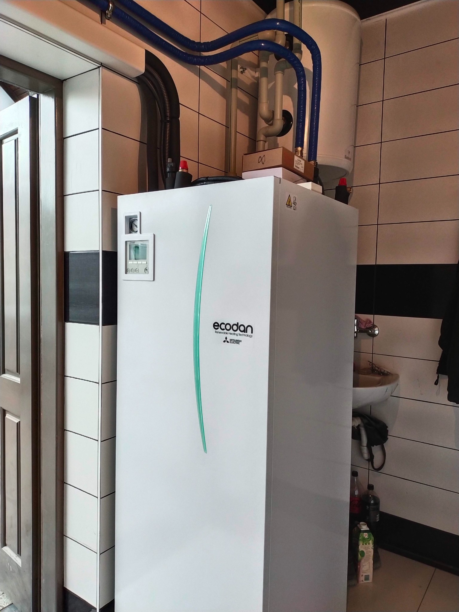 Biała jednostka wewnętrzna z niebieskimi rurami, marki Ecodan Mitsubishi, zamontowana w pomieszczeniu z biało-czarnymi płytkami, obok umywalki i drzwi.