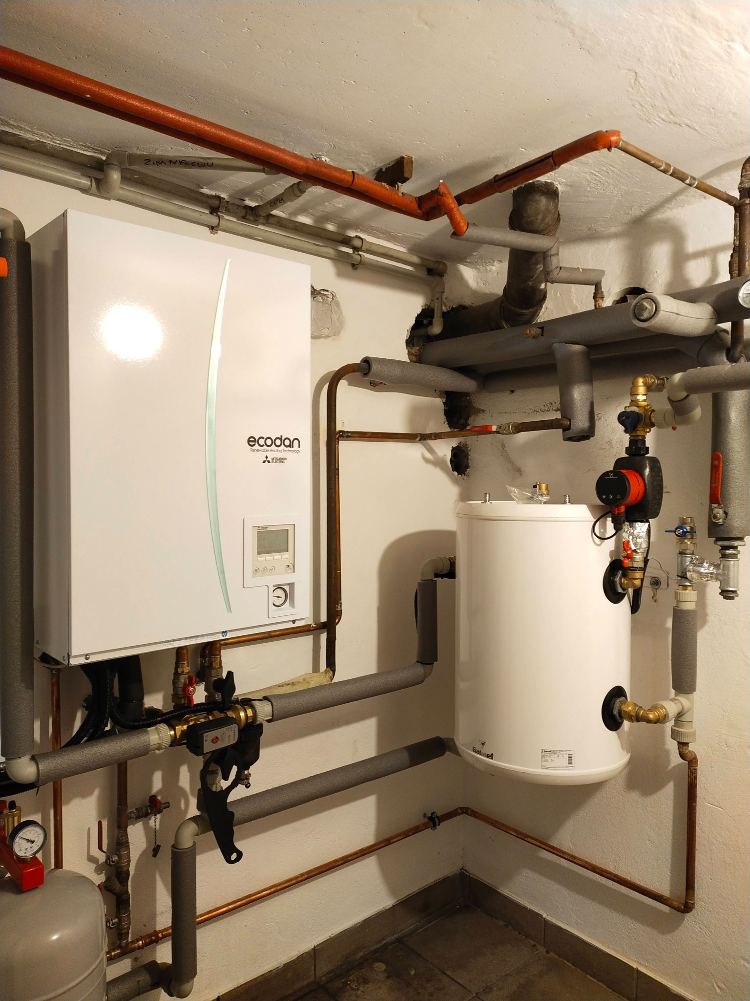 Biała jednostka wewnętrzna z pompą obiegową i zasobnikiem ciepłej wody użytkowej, instalacja grzewcza z rurami miedzianymi i izolacją, widoczne oznaczenia na rurach.