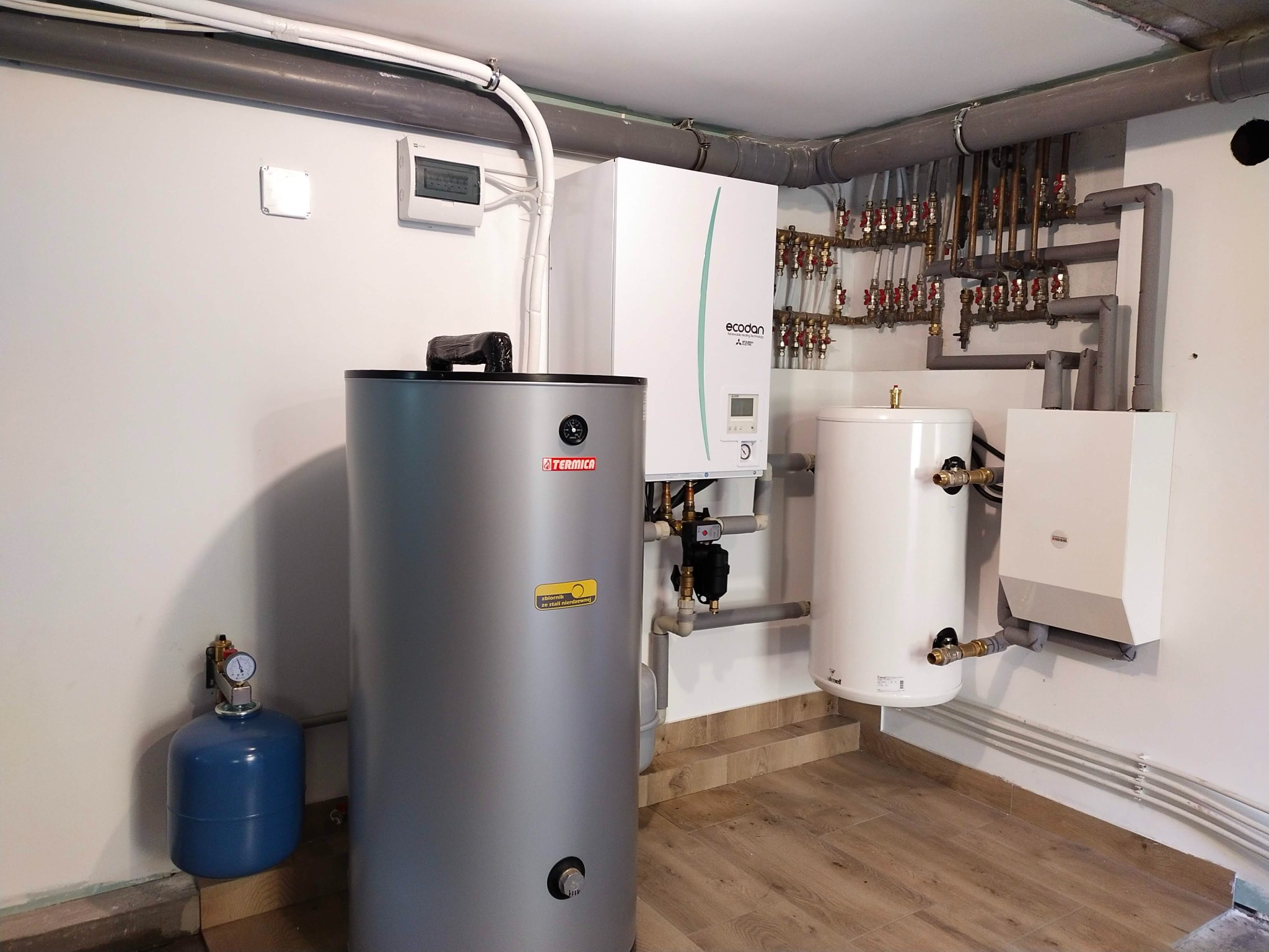 Kompletna instalacja grzewcza w kotłowni z szarym zbiornikiem Termica, białą jednostką Ecodan Mitsubishi, dodatkowym białym zbiornikiem i niebieskim naczyniem przeponowym, widoczne liczne rury...