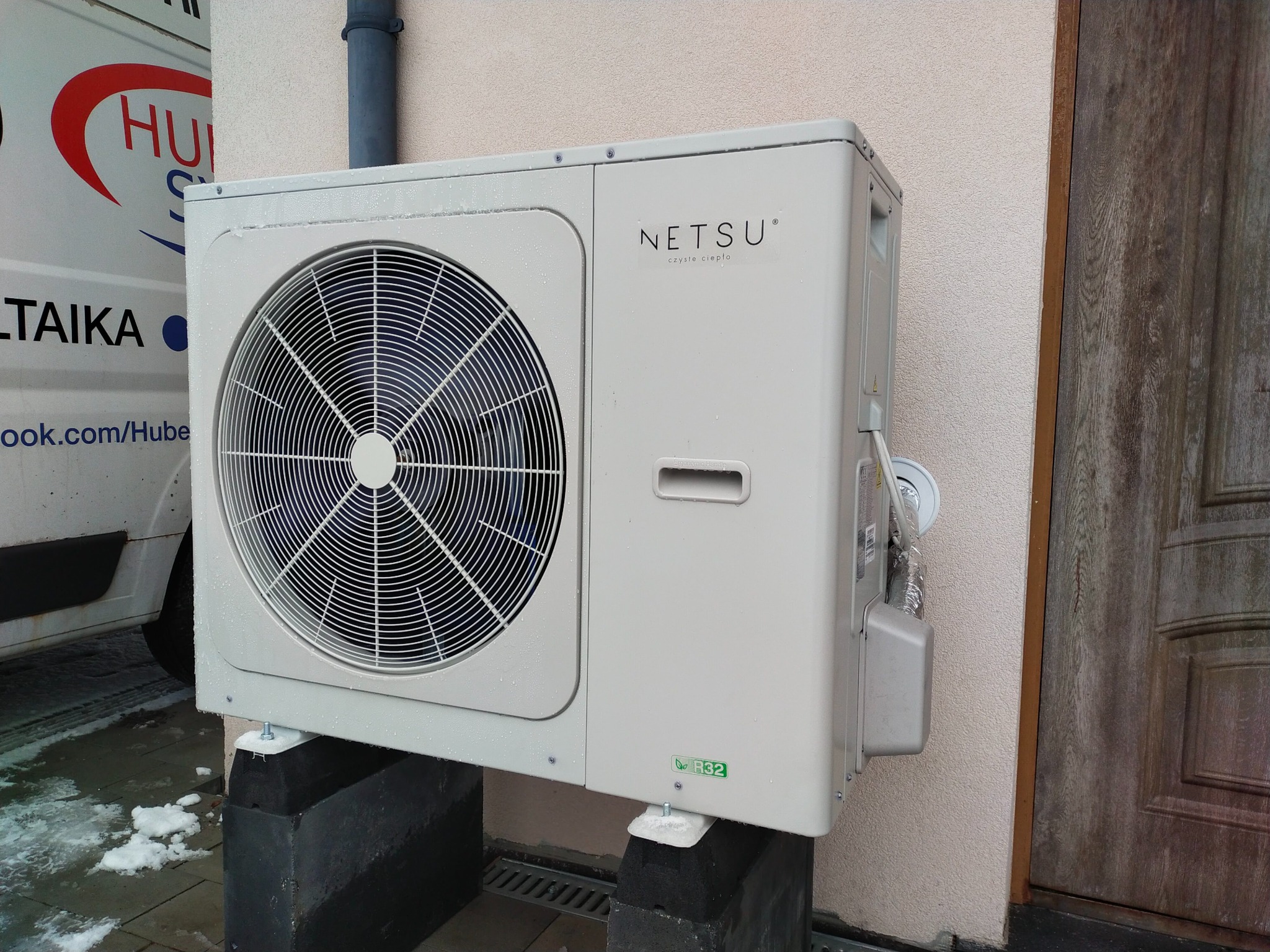 Zewnętrzna jednostka pompy ciepła marki Netsu zamontowana na betonowych wspornikach, z widocznymi śladami wilgoci i śniegiem w tle, obok drewnianych drzwi i częściowo widocznego samochodu firmy.