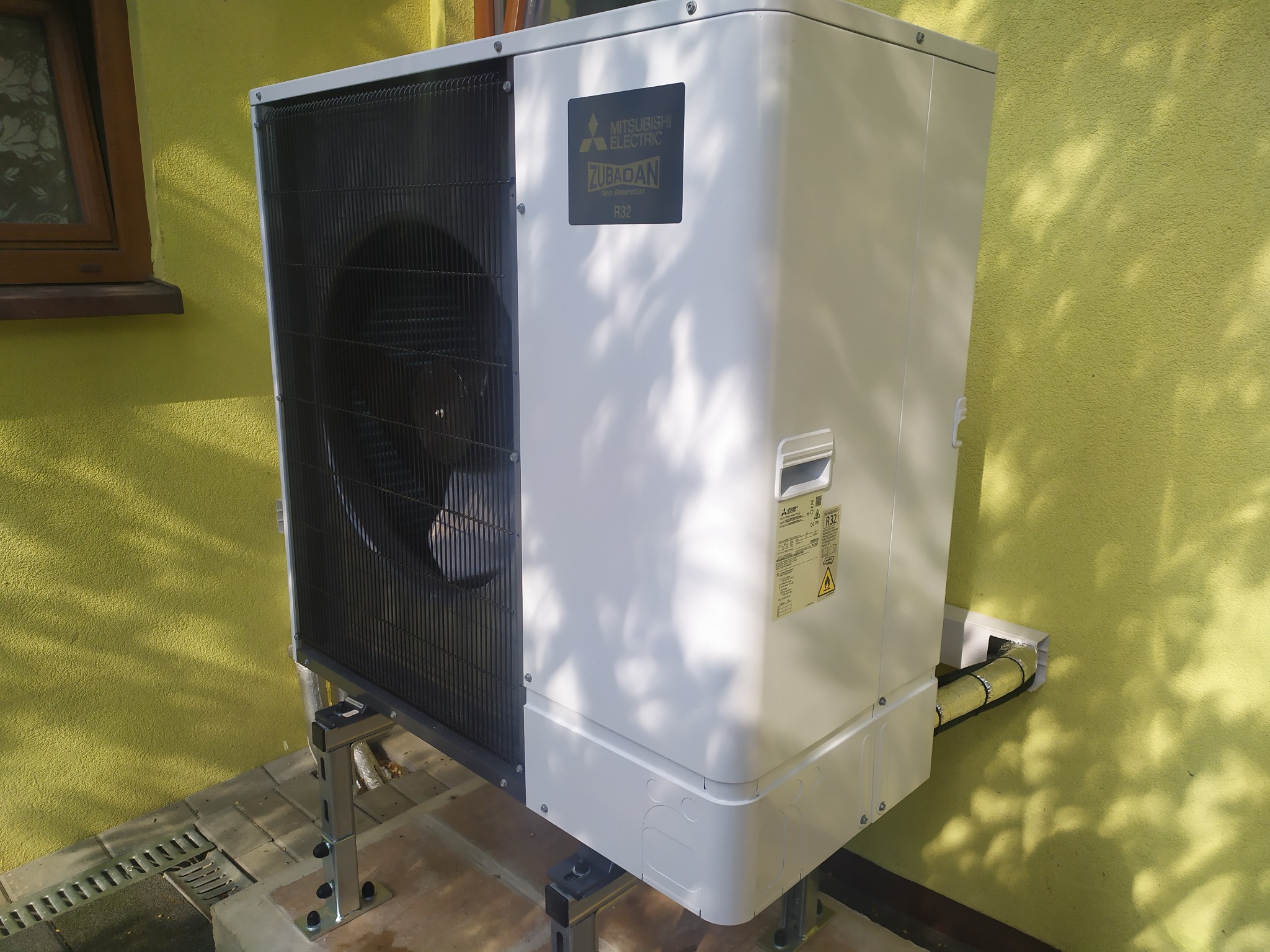 Zewnętrzna jednostka klimatyzacyjna Mitsubishi Zubadan R32 zamontowana na metalowej konstrukcji przy żółtej ścianie budynku, widoczne rury w izolacji prowadzące do ściany.