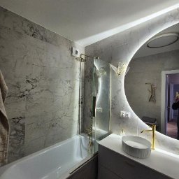 Kamar KAMIL ŚWICA - Nowoczesna łazienka z marmurowymi płytkami, podświetlanym lustrem i złotą armaturą, widoczne detale dekoracyjne w kształcie ptaków.