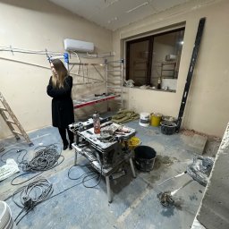 Kamar KAMIL ŚWICA - Wnętrze w trakcie remontu z drabiną, rusztowaniem, stołem roboczym z narzędziami, wiaderkami i stojącą kobietą w płaszczu.