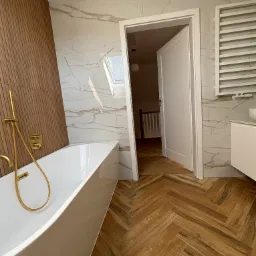 Nowoczesna łazienka z białą wanną wolnostojącą, złotą armaturą, marmurowymi płytkami na ścianach i drewnopodobną podłogą ułożoną w jodełkę; widok na otwarte drzwi do dalszej części domu.