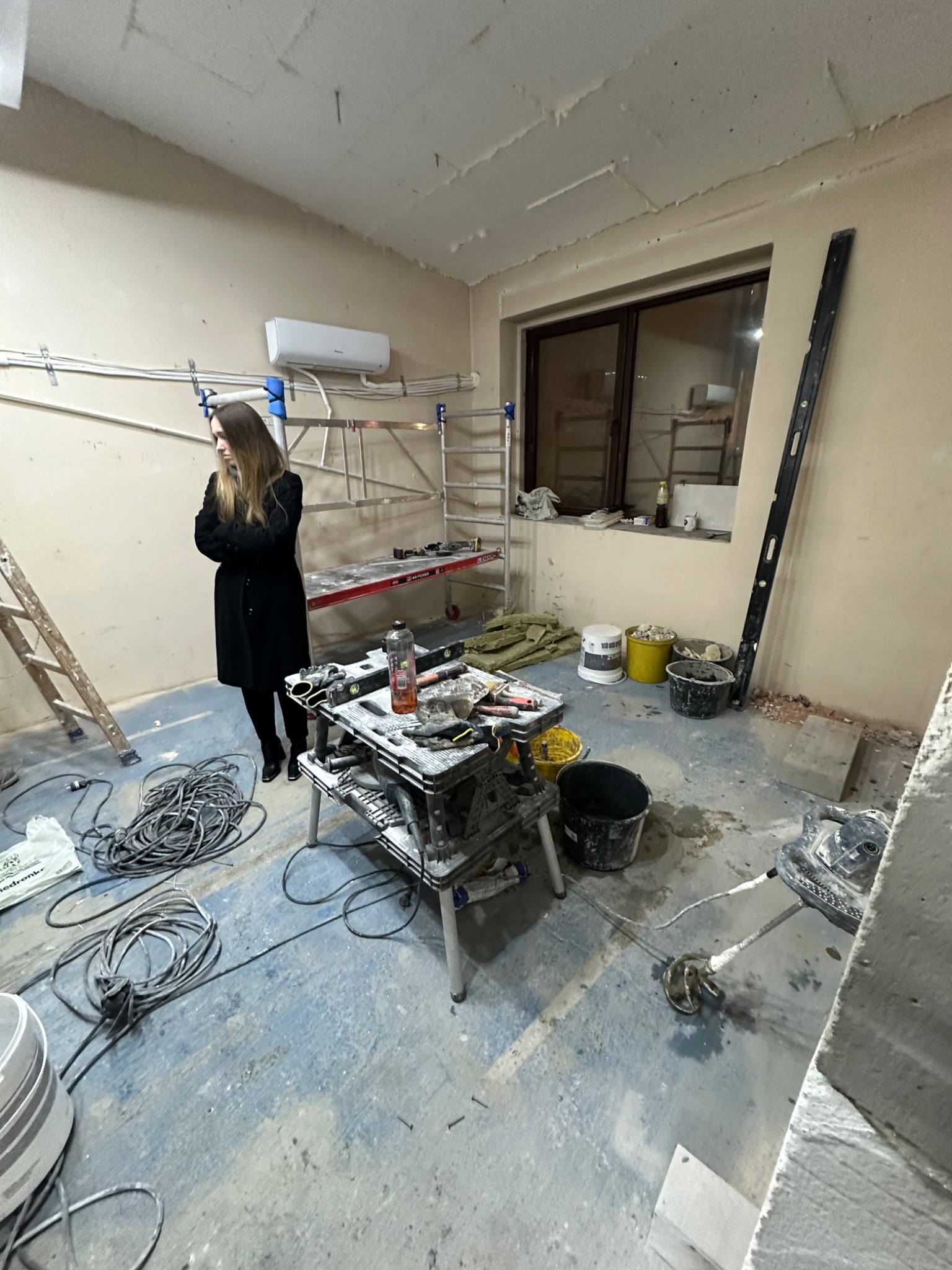 Wnętrze w trakcie remontu z drabiną, rusztowaniem, stołem roboczym z narzędziami, wiaderkami i stojącą kobietą w płaszczu.