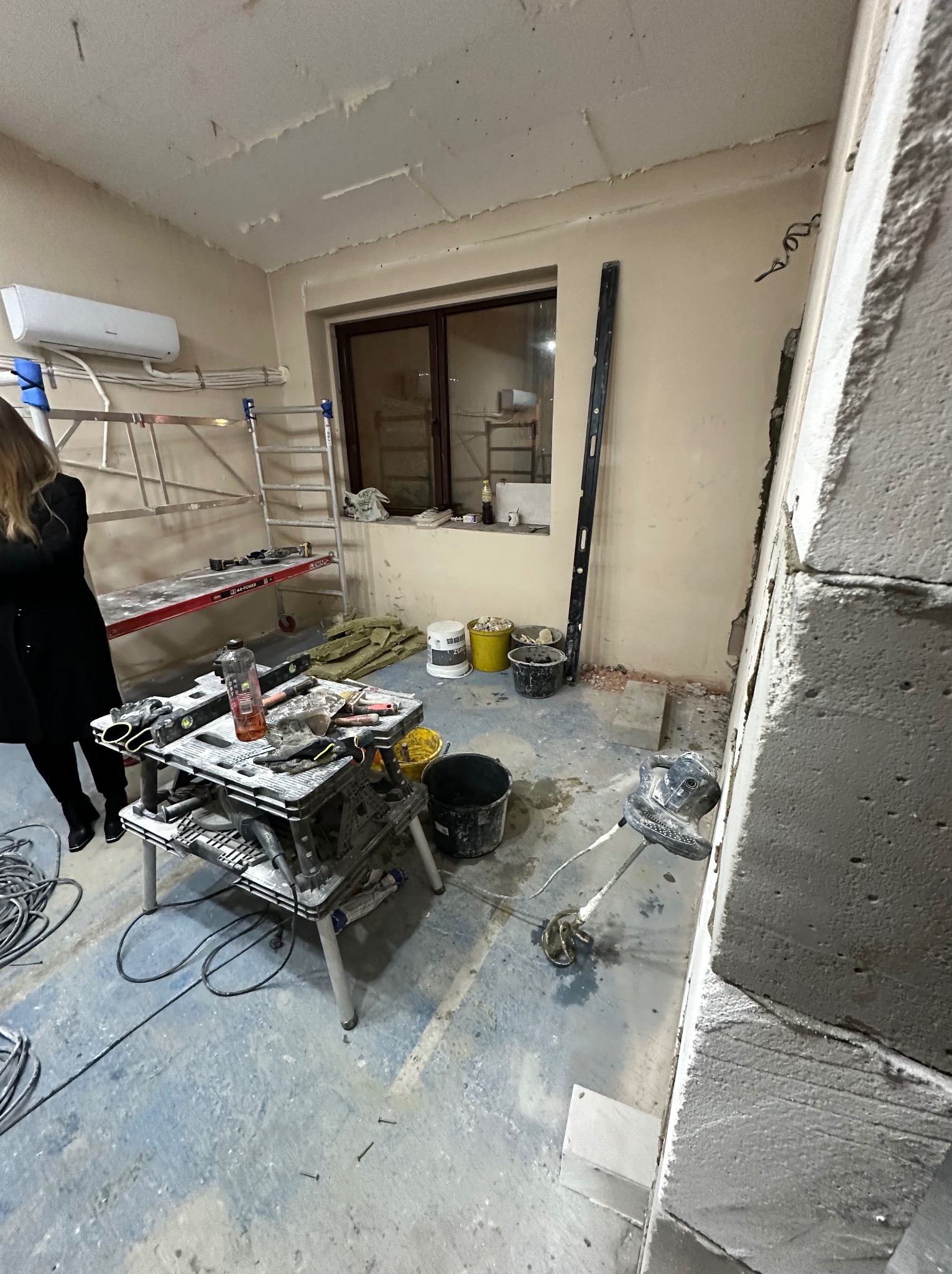 Wnętrze w trakcie prac budowlanych: widoczny stół roboczy z narzędziami, drabina, poziomica, szpachla elektryczna przy ścianie z betonu komórkowego, worki i pojemniki z materiałami budowlanymi...