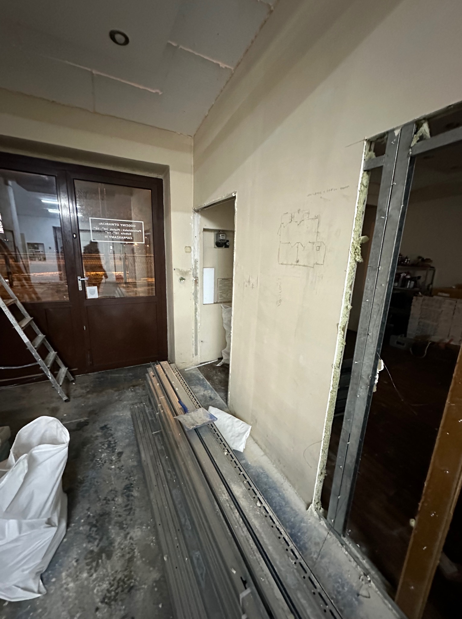 Wnętrze w trakcie prac wykończeniowych: widoczne profile metalowe, płyty gipsowo-kartonowe na ścianach z zaznaczonymi szkicami, drabina oparta o drzwi wejściowe, worek z materiałami budowlanymi.
