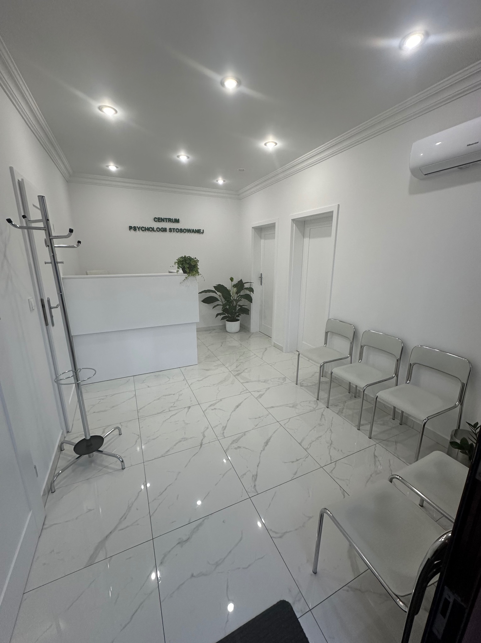 Jasne wnętrze poczekalni z białą recepcją, roślinami doniczkowymi i podłogą z płytek imitujących marmur, oświetlone punktowymi lampami sufitowymi i klimatyzatorem ściennym.