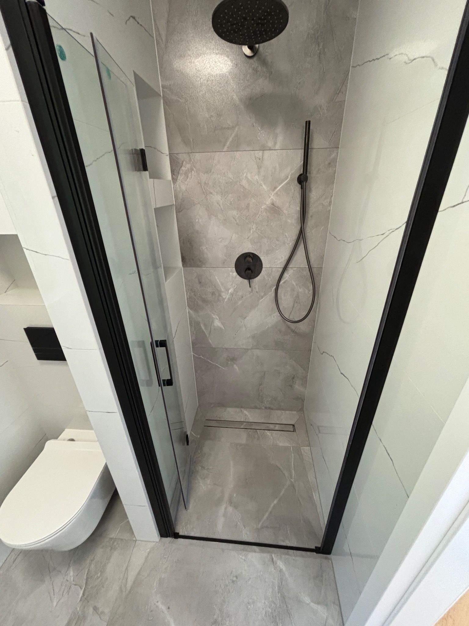 Nowoczesna łazienka z kabiną prysznicową typu walk-in z czarnymi ramami, deszczownicą i odpływem liniowym, wykończona płytkami imitującymi marmur.