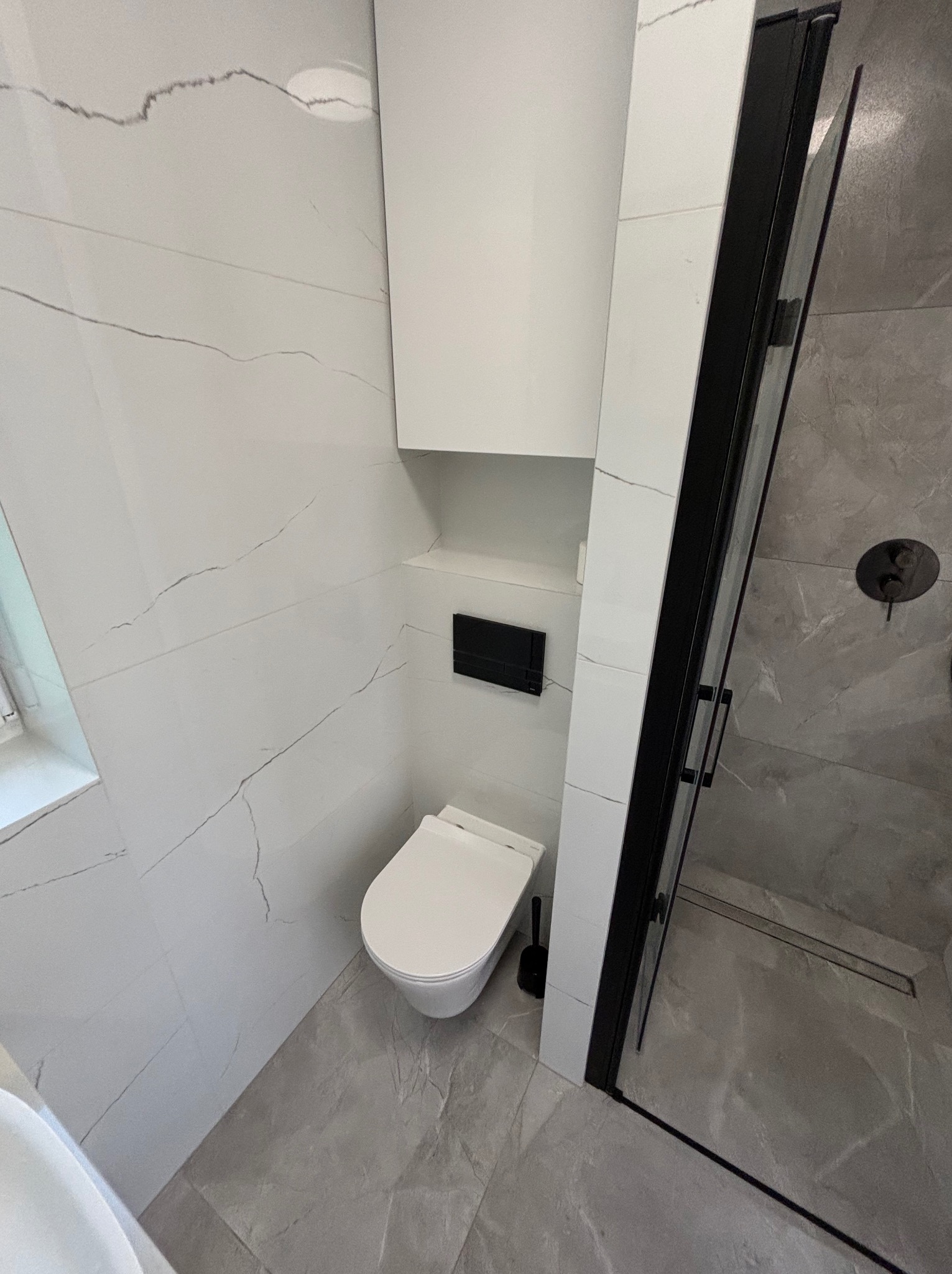 Nowoczesna łazienka z białymi płytkami imitującymi marmur, podwieszana toaleta, czarna armatura prysznicowa i kabina z czarnymi profilami.