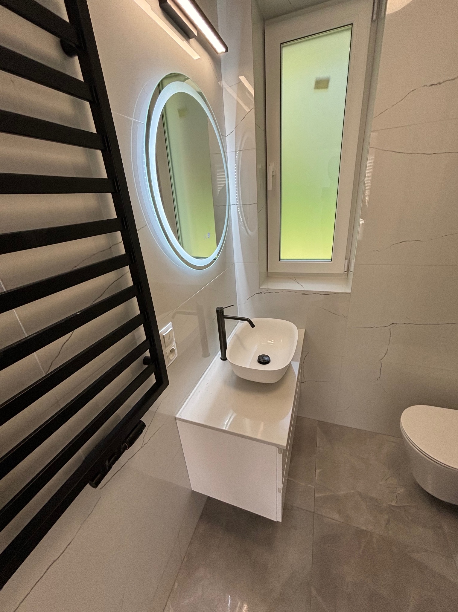 Nowoczesna łazienka z umywalką nablatową, podświetlanym okrągłym lustrem i czarnym grzejnikiem drabinkowym na tle białych płytek imitujących marmur.