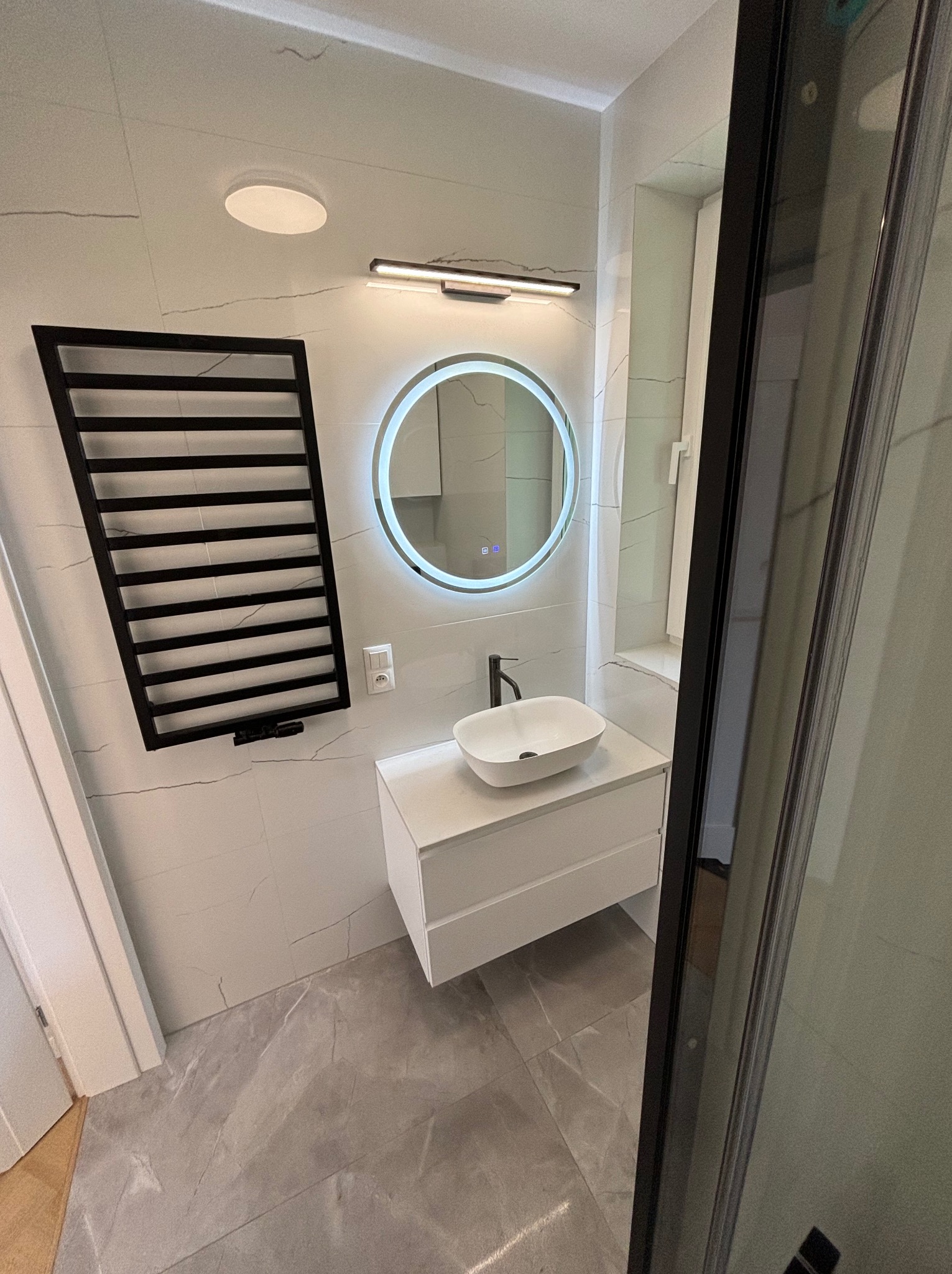Nowoczesna łazienka z białymi płytkami imitującymi marmur, okrągłym lustrem z podświetleniem LED, białą szafką pod umywalką i czarnym grzejnikiem drabinkowym.