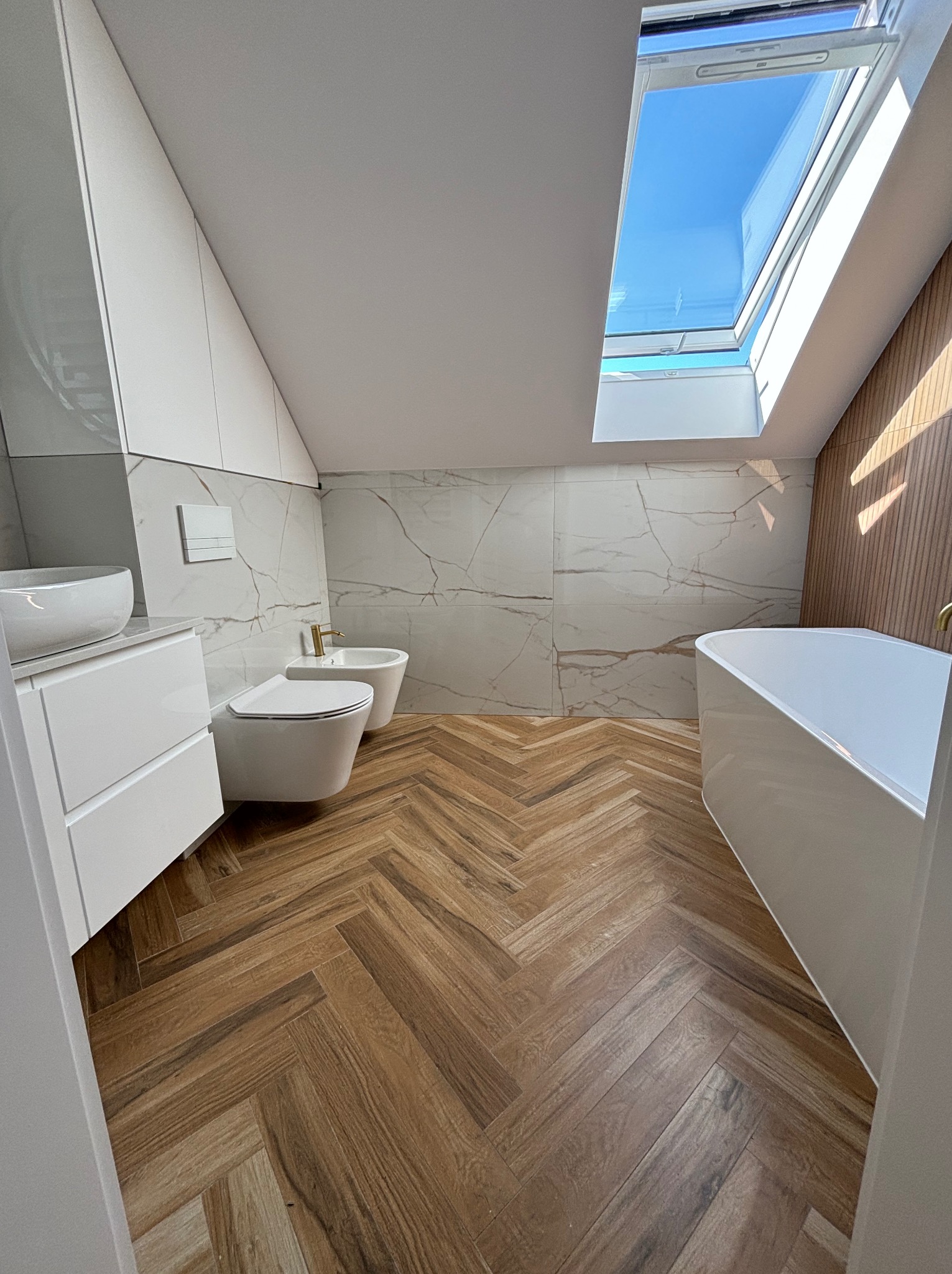 Nowoczesna łazienka na poddaszu z drewnopodobną podłogą ułożoną w jodełkę, białą armaturą, marmurowymi płytkami ściennymi i oknem dachowym wpuszczającym naturalne światło.
