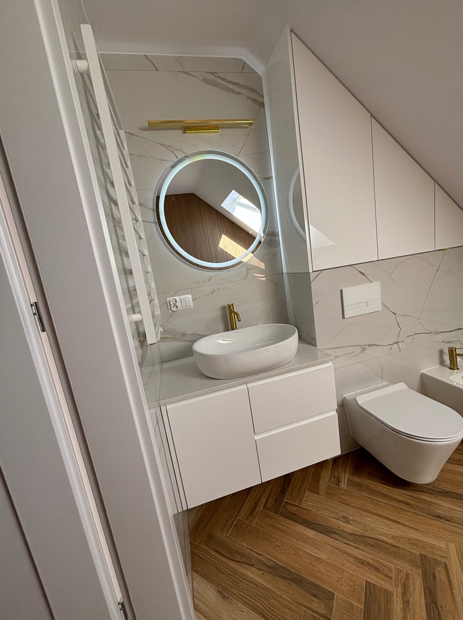 Nowoczesna łazienka na poddaszu z białą armaturą, okrągłym lustrem z podświetleniem LED, marmurowymi płytkami ściennymi i drewnianą podłogą ułożoną w jodełkę.