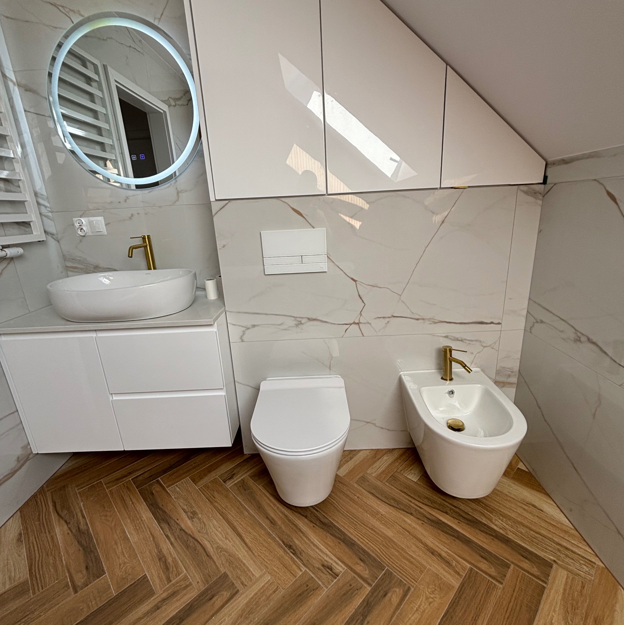Nowoczesna łazienka z białą armaturą, złotą baterią umywalkową i bidetową, marmurowymi płytkami ściennymi i podłogą w jodełkę z paneli drewnopodobnych, oświetlone lustro i białe szafki podwieszane.