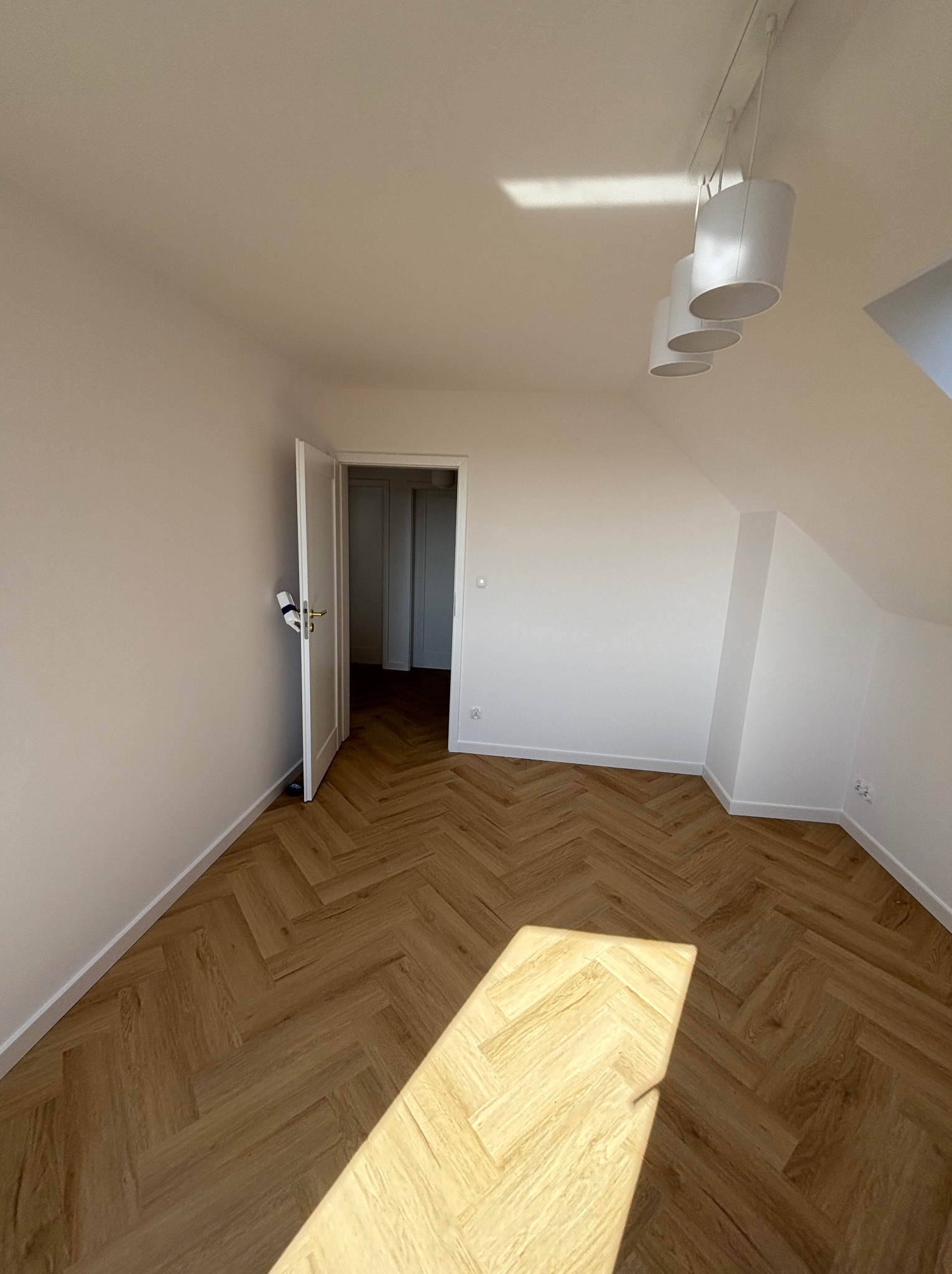 Pokój po remoncie z parkietem ułożonym w jodełkę, świeżo pomalowane ściany na biało, otwarte drzwi do sąsiedniego pomieszczenia, nowoczesne oświetlenie sufitowe z trzema cylindrycznymi lampami.