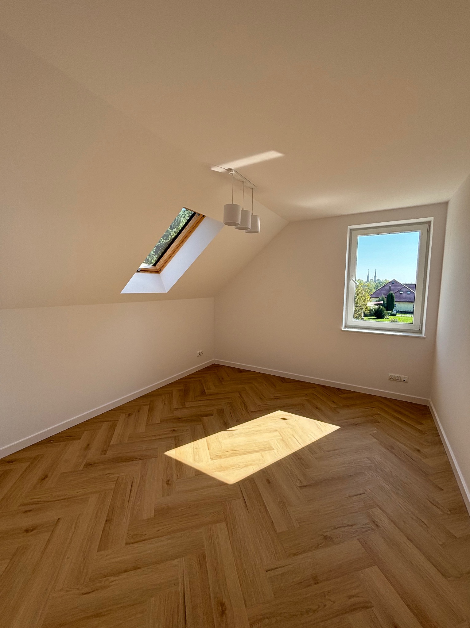 Puste, jasne poddasze z parkietem ułożonym w jodełkę, oświetlone przez okno dachowe i okno z widokiem na kościół w oddali.