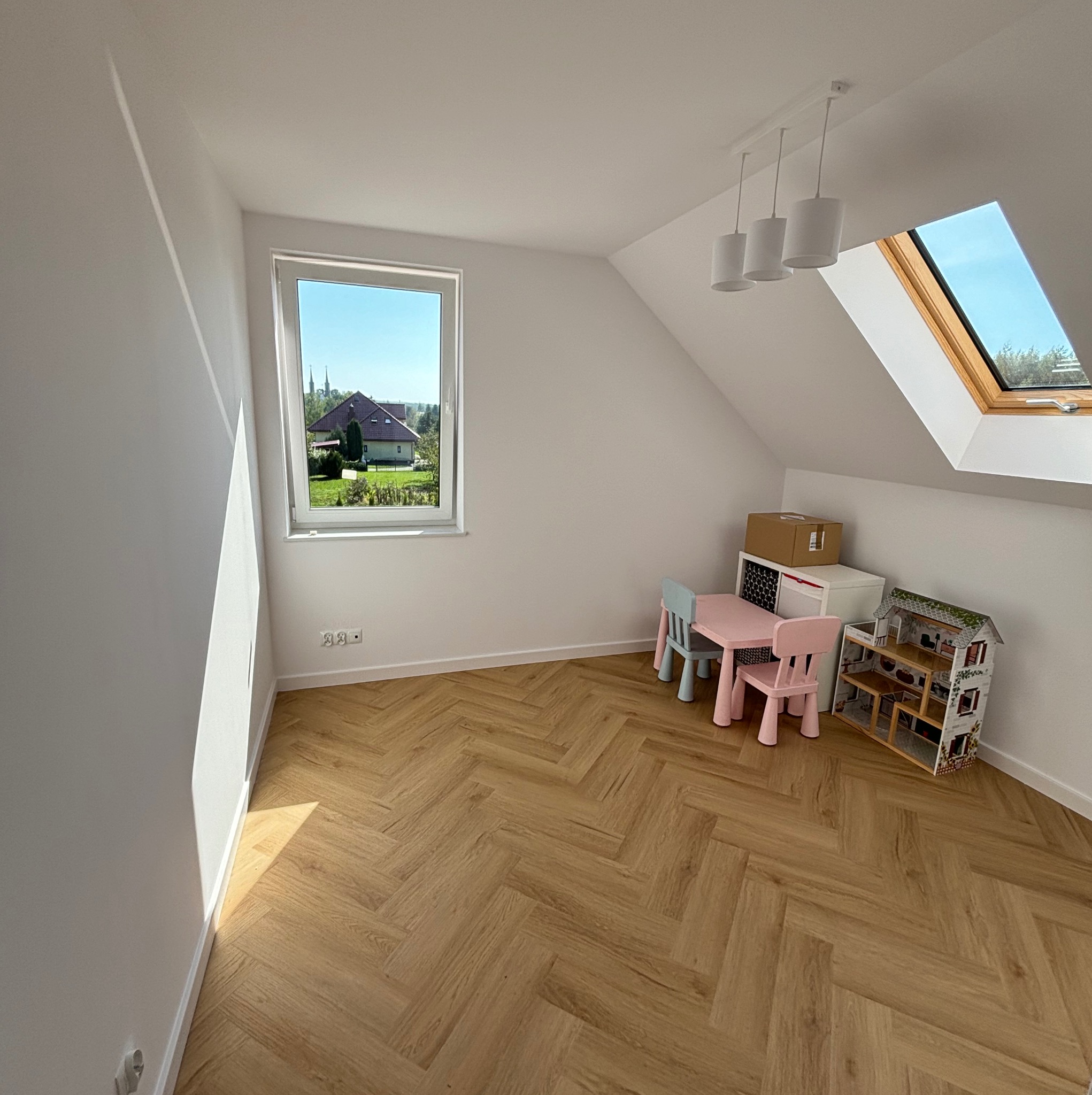 Jasne, nowocześnie wykończone poddasze z oknem dachowym i parkietem ułożonym we wzór jodełki francuskiej. Widoczne dziecięce mebelki i domek dla lalek.