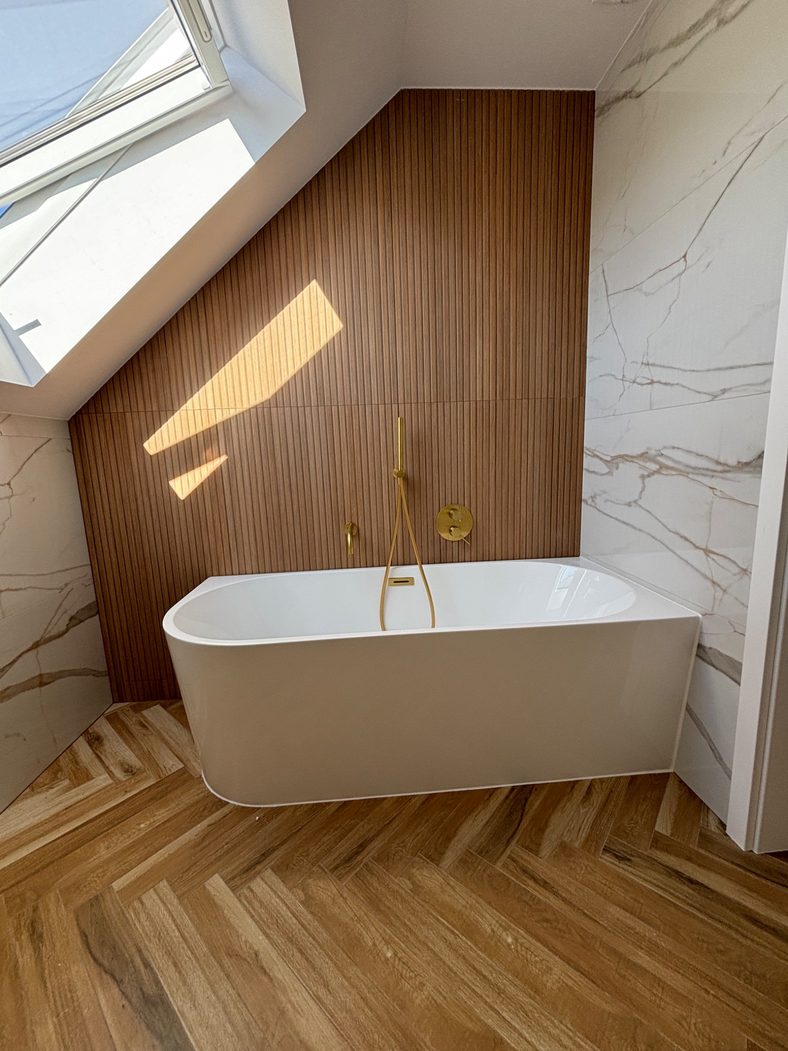 Biała wanna wolnostojąca z złotą armaturą na tle drewnianej ściany z pionowych paneli i marmurowych płytek, oświetlona naturalnym światłem z okna dachowego, na podłodze parkiet ułożony w jodełkę.