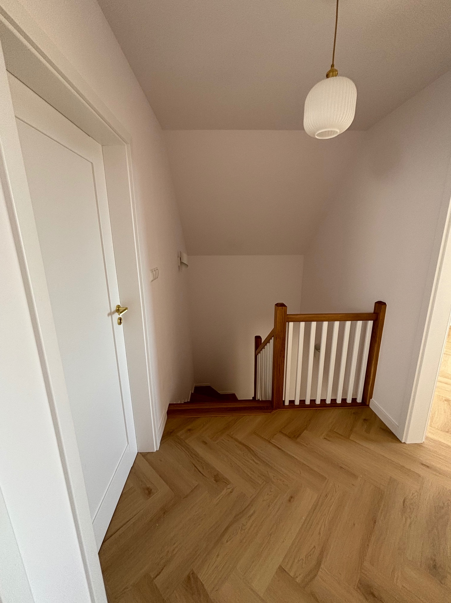 Korytarz z drewnianą podłogą ułożoną w jodełkę, białymi ścianami, drzwiami i schodami z drewnianą balustradą. W tle widoczna lampa sufitowa.