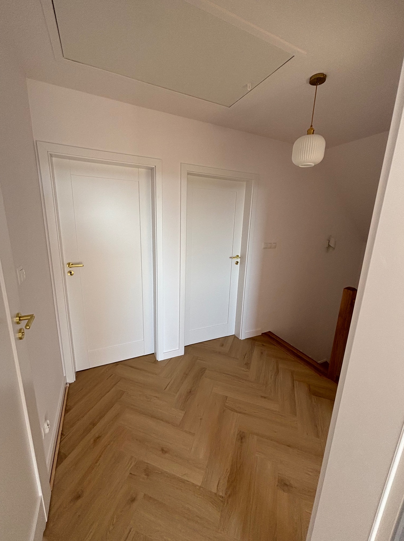 Korytarz z parkietem ułożonym w jodełkę, białymi drzwiami z mosiężnymi klamkami i wiszącą lampą z mlecznego szkła.