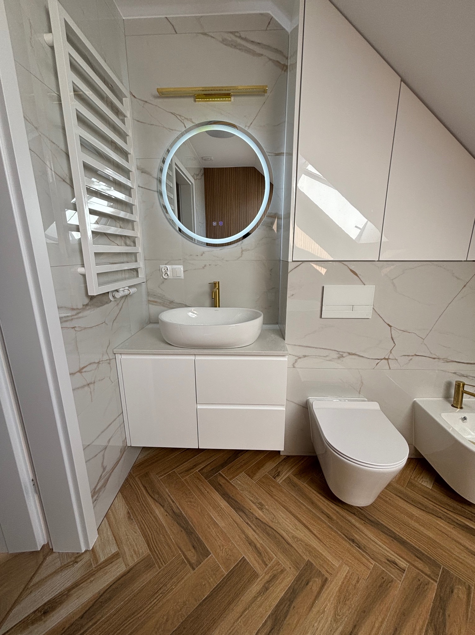 Nowoczesna łazienka na poddaszu z białymi meblami, umywalką nablatową i okrągłym lustrem z podświetleniem LED, wykończona płytkami imitującymi marmur i drewnopodobną podłogą ułożoną w jodełkę.