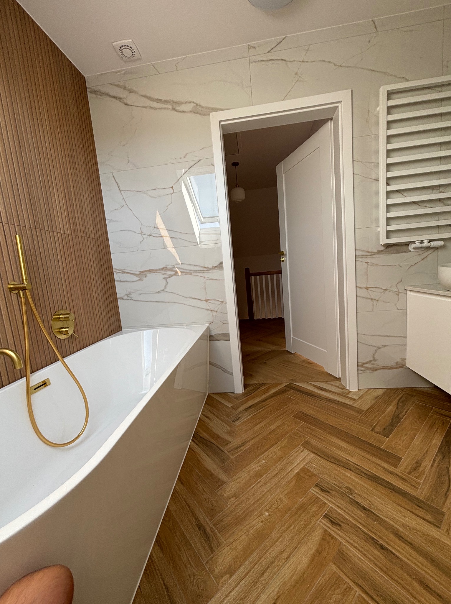 Nowoczesna łazienka z białą wanną wolnostojącą, złotą armaturą, marmurowymi płytkami na ścianach i drewnopodobną podłogą ułożoną w jodełkę; widok na otwarte drzwi do dalszej części domu.