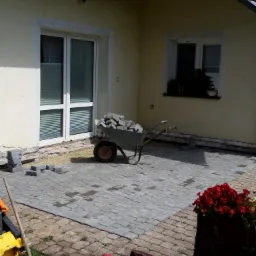 Fragment podwórka z częściowo ułożoną szarą kostką brukową, taczka wypełniona kostką, przygotowane podłoże, narzędzia i czerwone kwiaty w donicy.