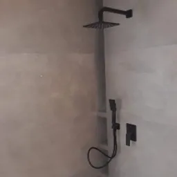 Minimalistyczna łazienka z czarną armaturą: deszczownica, słuchawka prysznicowa i bateria, na tle ściany wykończonej gładzią w odcieniach szarości.