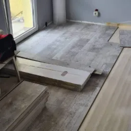 Układanie paneli podłogowych w pokoju z szarą ścianą, widoczna paczka paneli i podkład, częściowo ułożona podłoga z panelami imitującymi drewno.