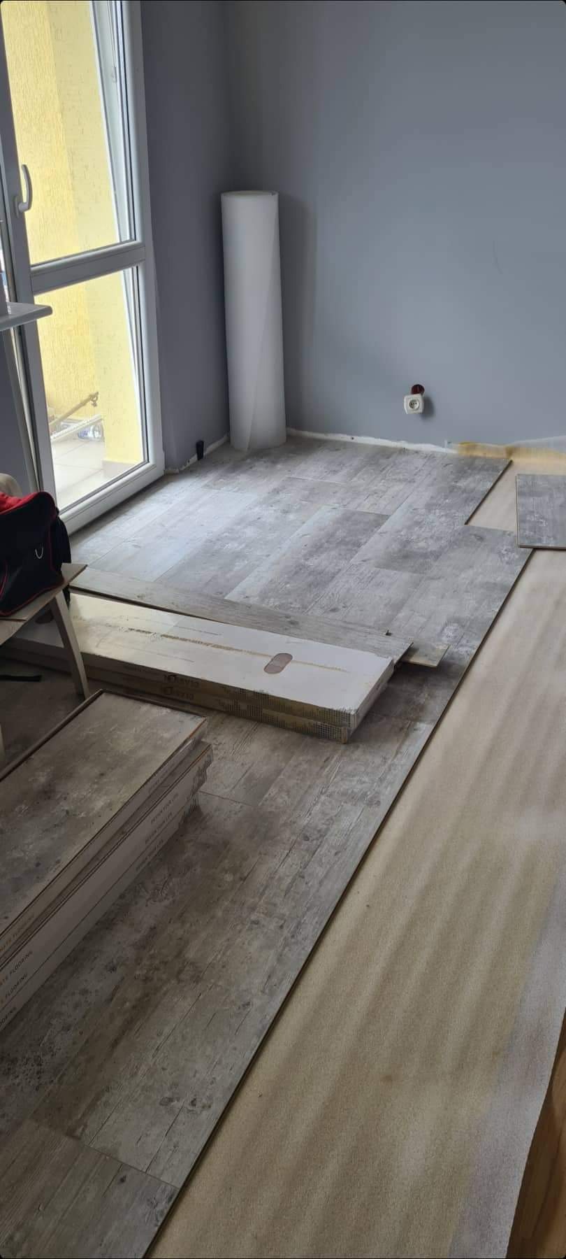 Układanie paneli podłogowych w pokoju z szarą ścianą, widoczna paczka paneli i podkład, częściowo ułożona podłoga z panelami imitującymi drewno.
