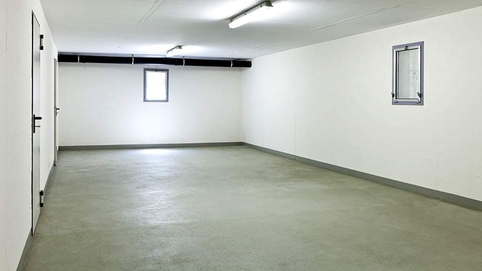 Wnętrze pustego, odnowionego pomieszczenia z betonową podłogą, białymi ścianami, dwojgiem drzwi i małym oknem z szarą ramą. Widoczne rury pod sufitem i listwy przypodłogowe.
