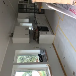 Wnętrze kuchni w trakcie montażu: białe szafki kuchenne, okna z widokiem na zieleń, podłoga zabezpieczona taśmą malarską, widoczne kartony z wyposażeniem i narzędziami.