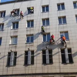 Grupa pracowników w uprzężach linowych, z wiadrami i narzędziami, zawieszona na elewacji budynku z licznymi oknami; w tle widoczny miejski autobus i sygnalizacja świetlna.