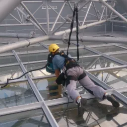 Pracownik zabezpieczony linami, w kasku, czyści szklany dach galerii handlowej za pomocą specjalistycznego sprzętu zasilanego kablem.