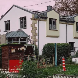 Dwupiętrowy dom z białą elewacją, elementami wykończeniowymi w odcieniach beżu i szarości, widoczne panele słoneczne na dachu, otoczony zielenią, z małą czerwoną budką i ozdobną latarnią morską...