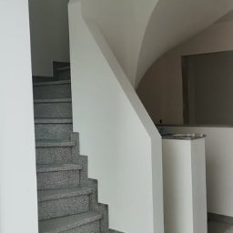 TomaszMajewski - Klatka schodowa z szarymi, kamiennymi schodami i świeżo pomalowanymi na biało ścianami. Widoczne przejście na wyższy poziom z łukowym elementem. Fragment podłogi z widocznymi zabrudzeniami.