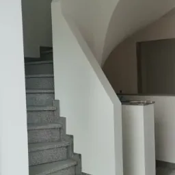 Klatka schodowa z szarymi, kamiennymi schodami i świeżo pomalowanymi na biało ścianami. Widoczne przejście na wyższy poziom z łukowym elementem. Fragment podłogi z widocznymi zabrudzeniami.