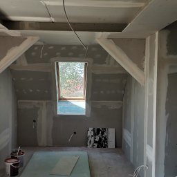 TomaszMajewski - Poddasze w trakcie prac wykończeniowych, widoczne płyty kartonowo-gipsowe na ścianach i suficie, okno dachowe oraz elementy konstrukcyjne pod skosami, częściowo zaszpachlowane łączenia, na podłodze...