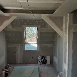 Poddasze w trakcie prac wykończeniowych, widoczne płyty kartonowo-gipsowe na ścianach i suficie, okno dachowe oraz elementy konstrukcyjne pod skosami, częściowo zaszpachlowane łączenia, na podłodze...